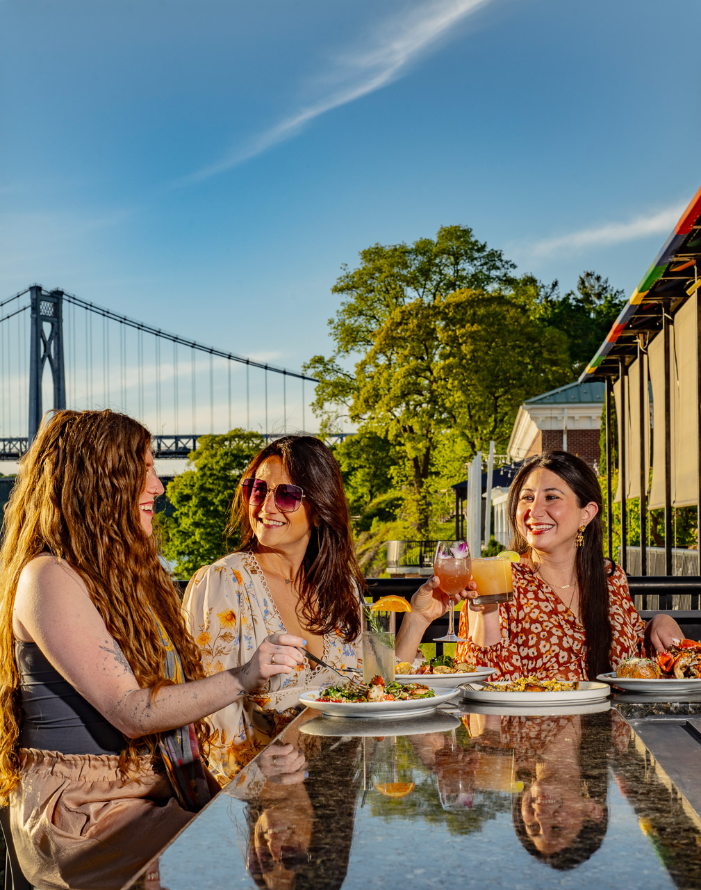 Shadows-on-the-Hudson-Summer2026-cocktails-outdoor-bar-models-10 Guests dining outdoors with the Mid-Hudson Bridge in the background.
